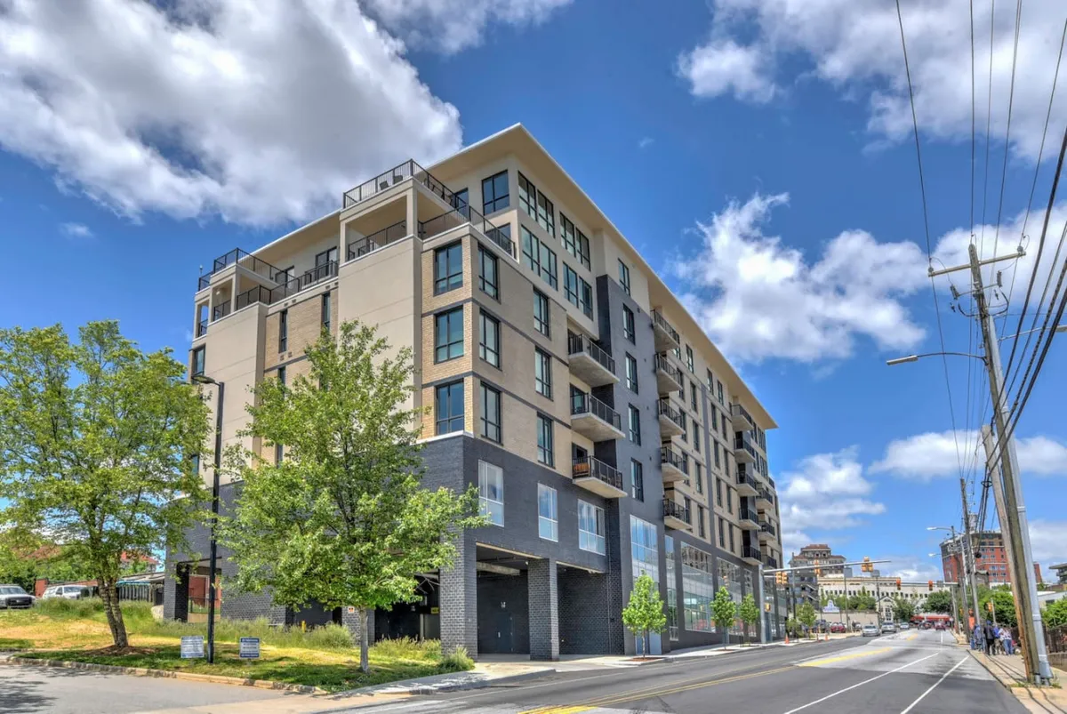 This image features a contemporary building with prominent balconies outlined by glass railings. The lush greenery in the foreground adds a touch of tranquility, while the city skyline is visible from the balconies, suggesting expansive urban views and an inviting residential atmosphere.
