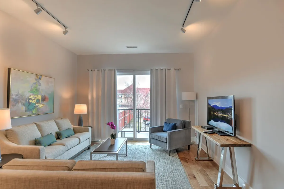 This image features a cozy, light-filled living room with a plush sofa, a coffee table, modern artwork, and a TV. Large balcony doors allow natural light to flow in, and glimpses of the city can be seen beyond. The setup invites relaxation and socializing, with comfortable seating and an open, airy feel.