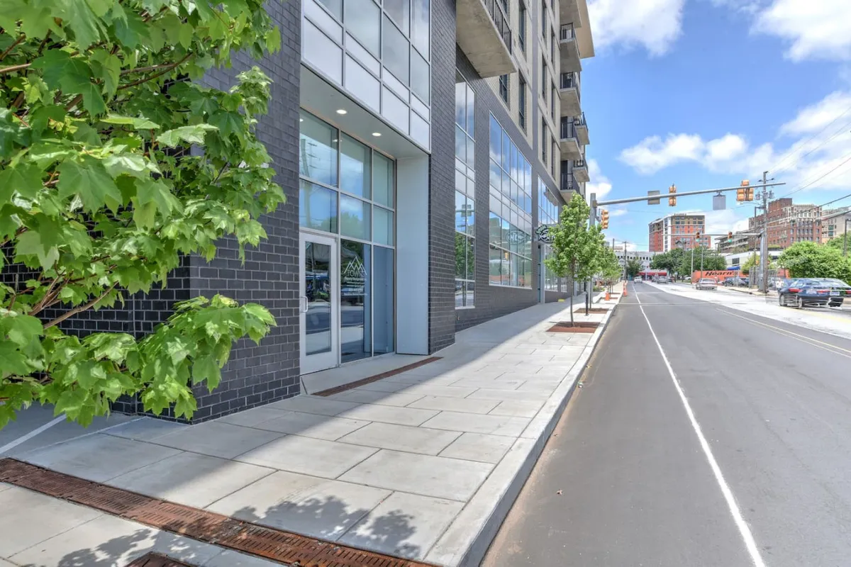 A lively urban streetscape featuring a contemporary building with large glass windows, mature street trees, and a wide, welcoming sidewalk. People can be seen walking or socializing, capturing the buzz of city life. The scene hints at nearby cafés, shops, and local events, inviting exploration beyond the front door.