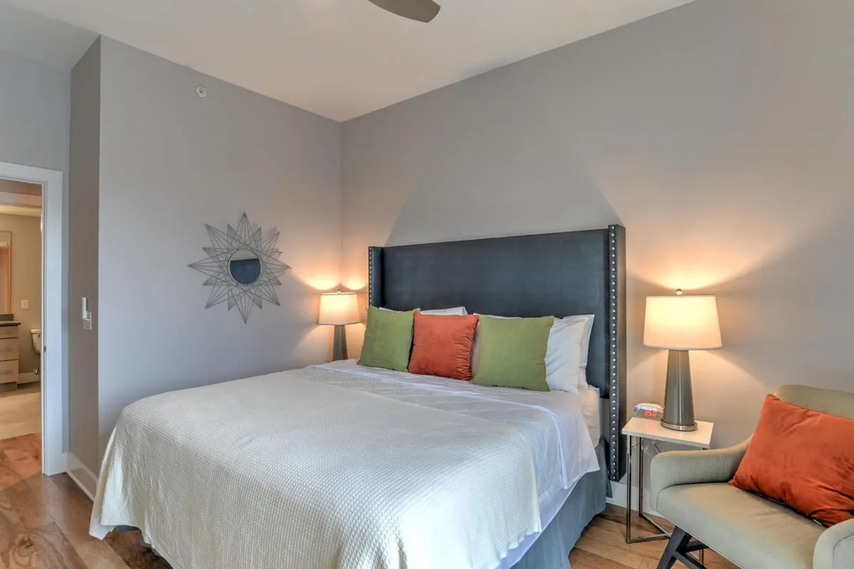 This image presents a welcoming bedroom with a neatly made bed, accent pillows, a mirror, table lamps, and a cozy armchair in the corner. The room is filled with natural light, enhancing the inviting mood, while practical touches like the armchair and mirror add to the feeling of a personalized retreat.