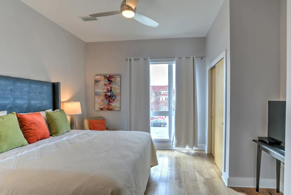 This photo highlights a cozy bed with colorful pillows, tasteful artwork, and a window allowing natural light to brighten the bedroom. The setting feels tranquil and inviting, ideal for unwinding after a day out or for peaceful mornings in.