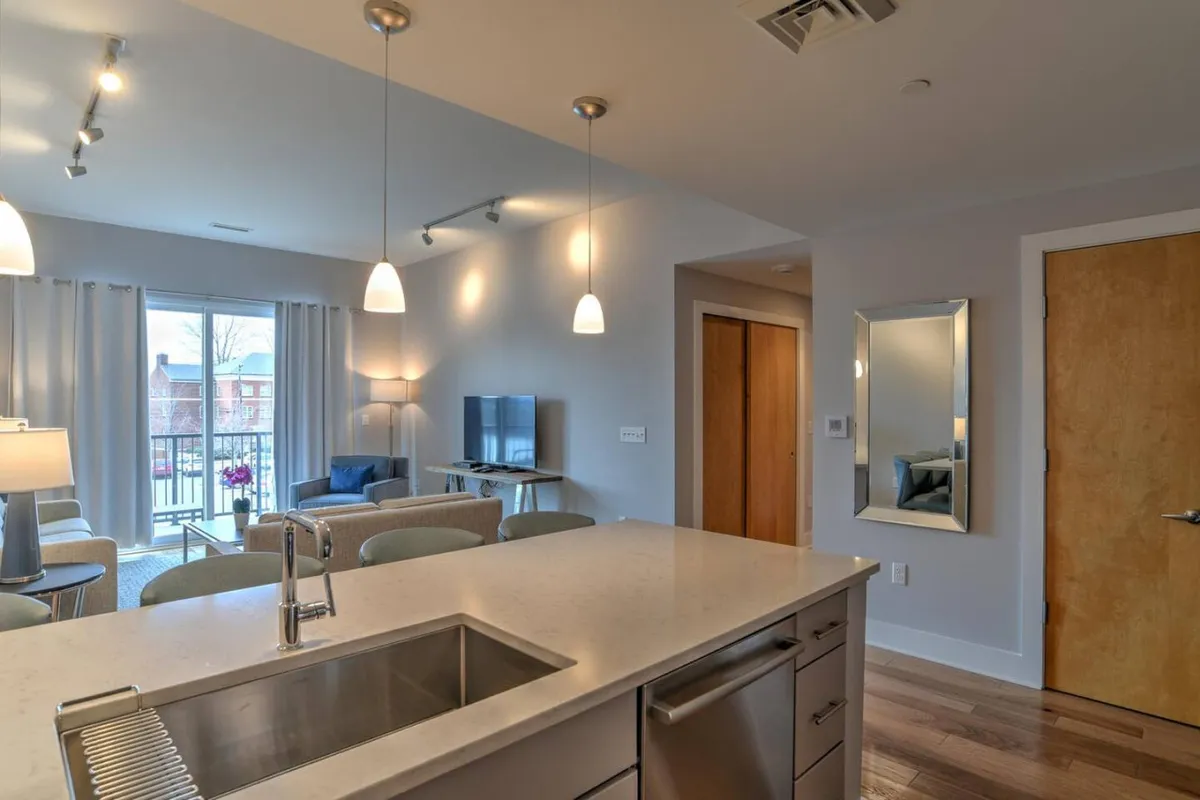 A wide-angle view showing the open-plan layout that seamlessly connects the living area with the kitchen. The kitchen island, visible window view, and adjacent living space convey a sense of flow and modern design, with ample room for both entertaining and everyday living.