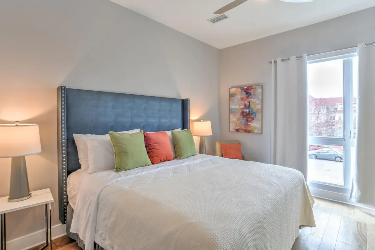 This image features a king-sized bed adorned with decorative pillows, a bedside table with a lamp, artwork on the wall, and a large window that fills the room with natural light. The atmosphere is serene and cozy, with a restful ambiance that invites relaxation and comfort.